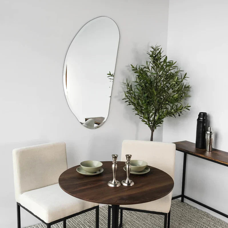 Artificial tree in the corner of a white room with an asymmetrical mirror, small brown wood bistro table and two off-white upholstered chairs beside small brown wood console table