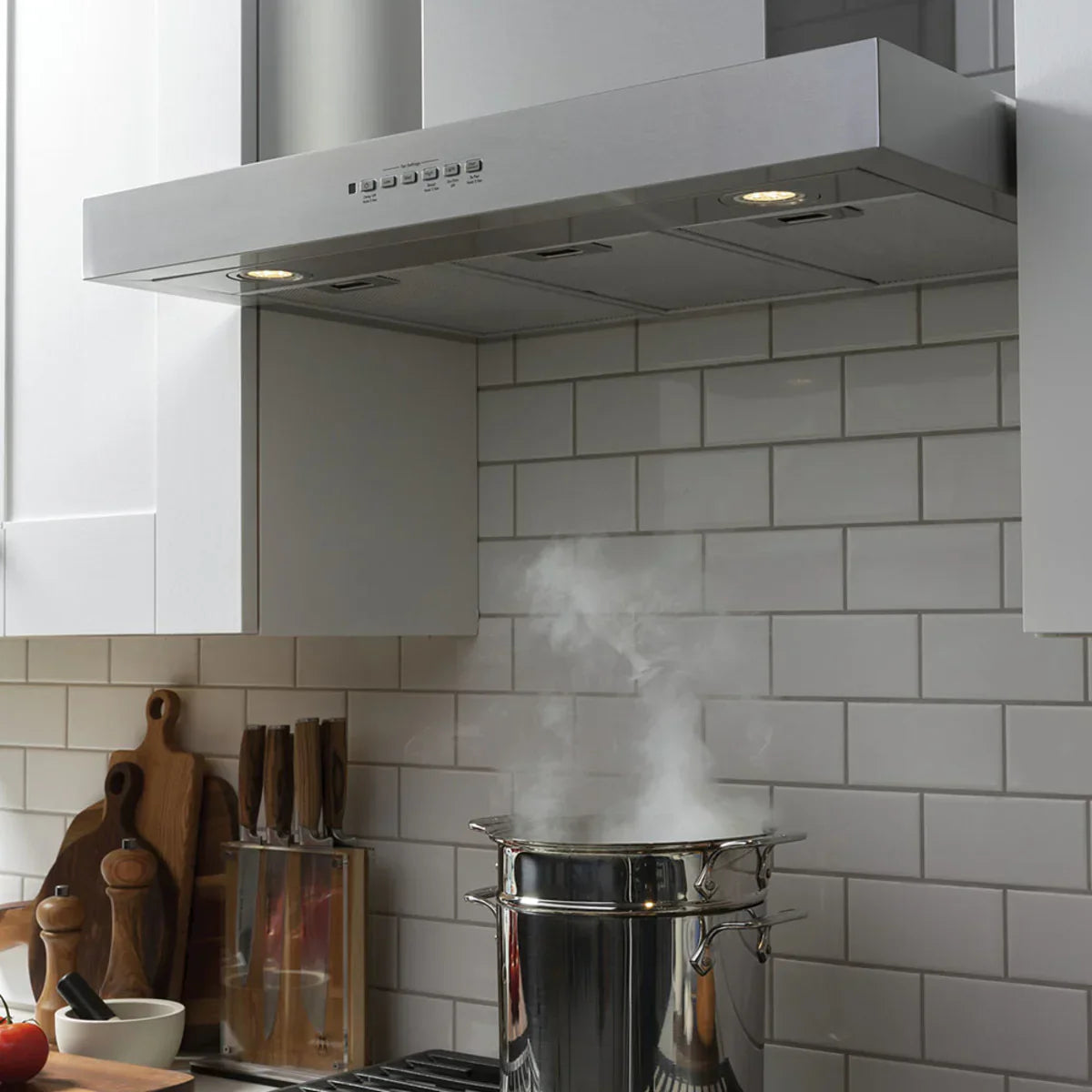 White kitchen with stainless steel wall mount range hood with lights over a pot of steaming water, and kitchen cooking utensils on the countertop beside it